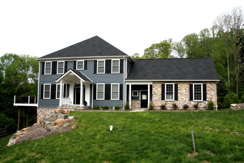 Front exterior of newly constructed house with teal siding, white trim, black shutters, gray roofing, and a stone garage.
