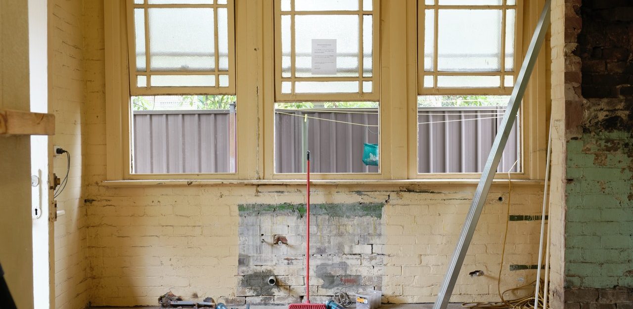 A red broom standing up against one of three windows in an unfinished room