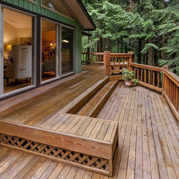 Large wraparound brown wooden back deck with built in seating and rails attached to a green house with large windows with large trees in the background.