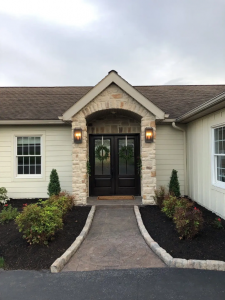 Front entrance of house after ranch style house modernization with new light stone veneer, two entry way lights, a path leading to the covered entrance, and flower beds on either side of the path with mulch and shrubs.